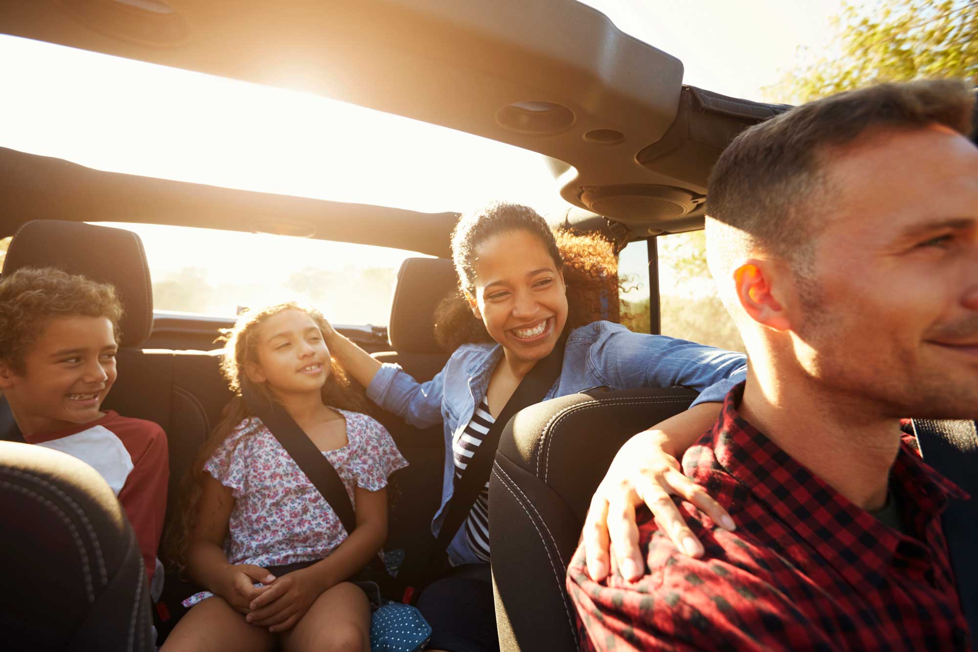 A father drives the car while the mother sits in teh backseat with 2 children, all smiling.